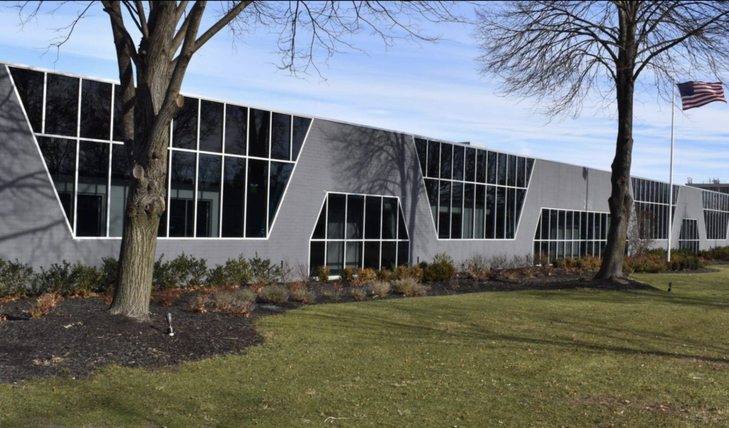 A modern building with irregularly shaped windows, gray walls, and an American flag on a pole in front, surrounded by grass, trees, and shrubs.